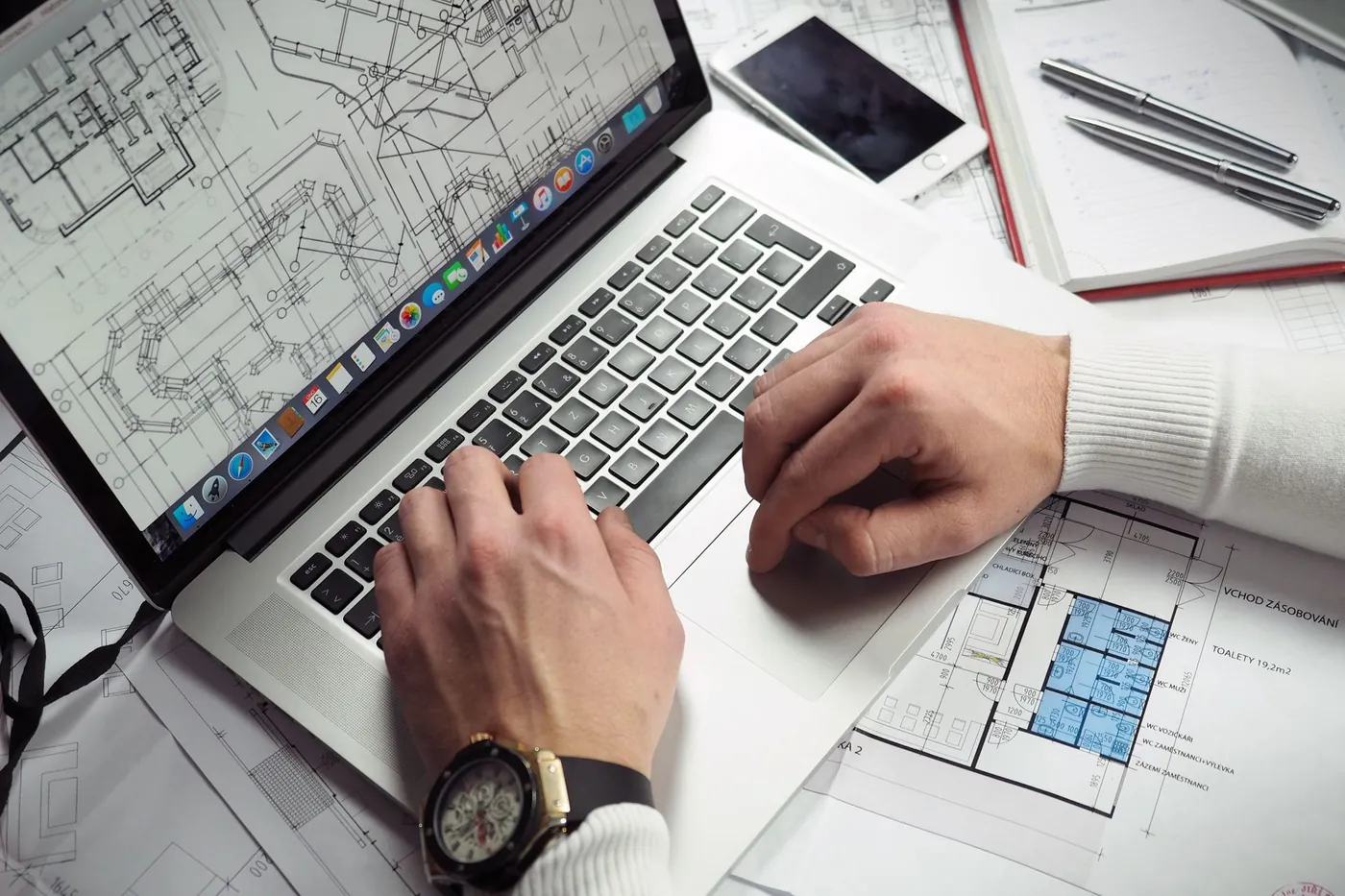 Mani al lavoro su un portatile, con le planimetrie di un progetto sul tavolo